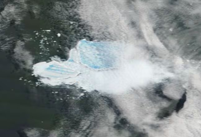 Immagine satellitare granulosa di un iceberg blu e bianco che si spezza sull'acqua scura, con nuvole bianche sopra di esso. 
