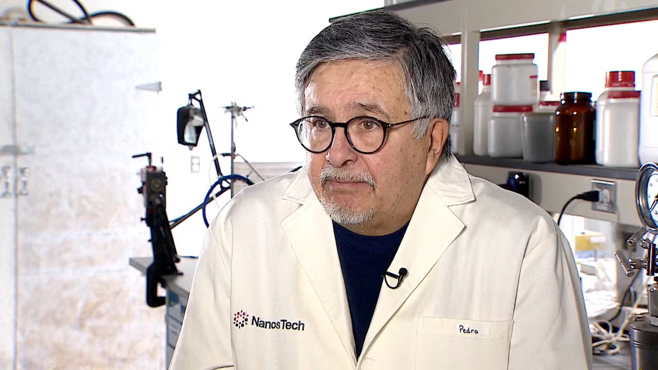 Un uomo in camice bianco viene intervistato con apparecchiature scientifiche sullo sfondo.