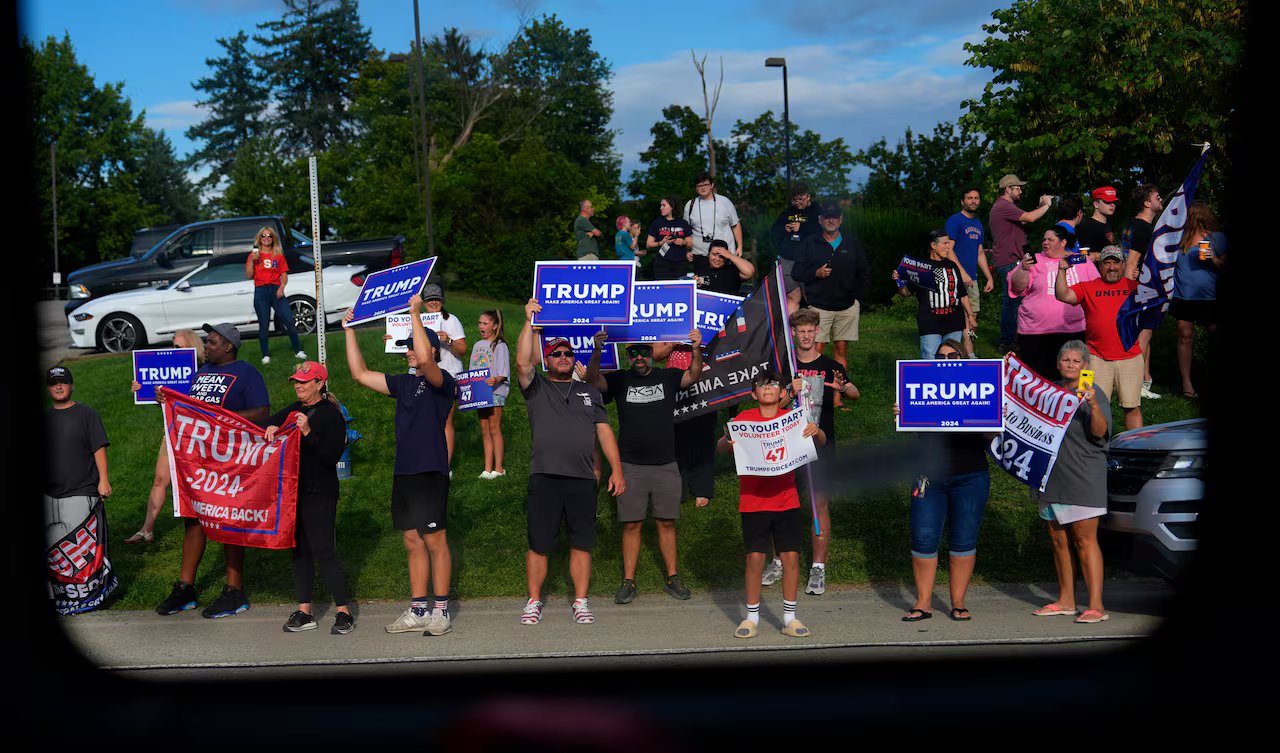 Attraverso il finestrino dell’autobus si possono vedere più di 20 persone in piedi sul bordo della strada, molte delle quali portano cartelli elettorali di Trump. 