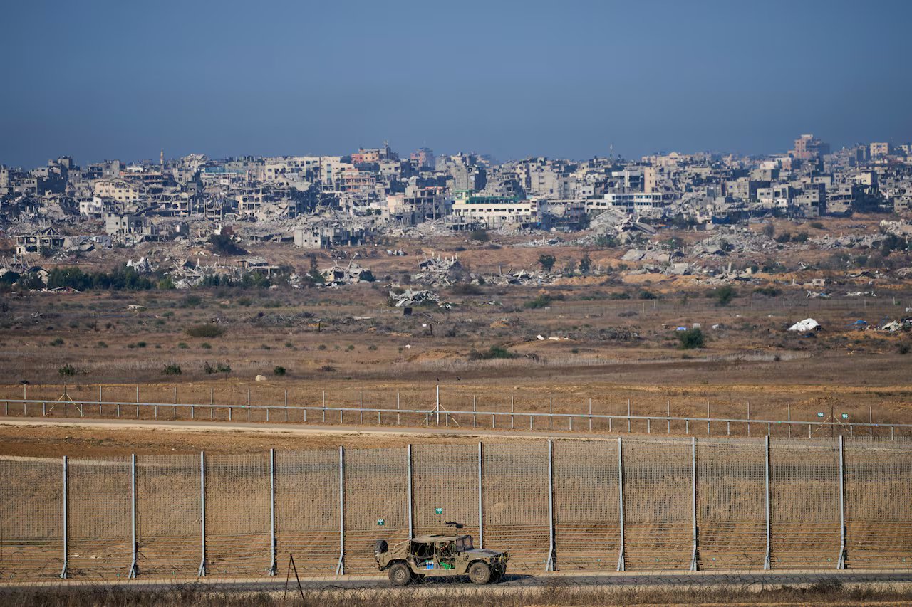 Un veicolo militare attraversa la recinzione di confine mentre dietro di essa compaiono edifici distrutti.