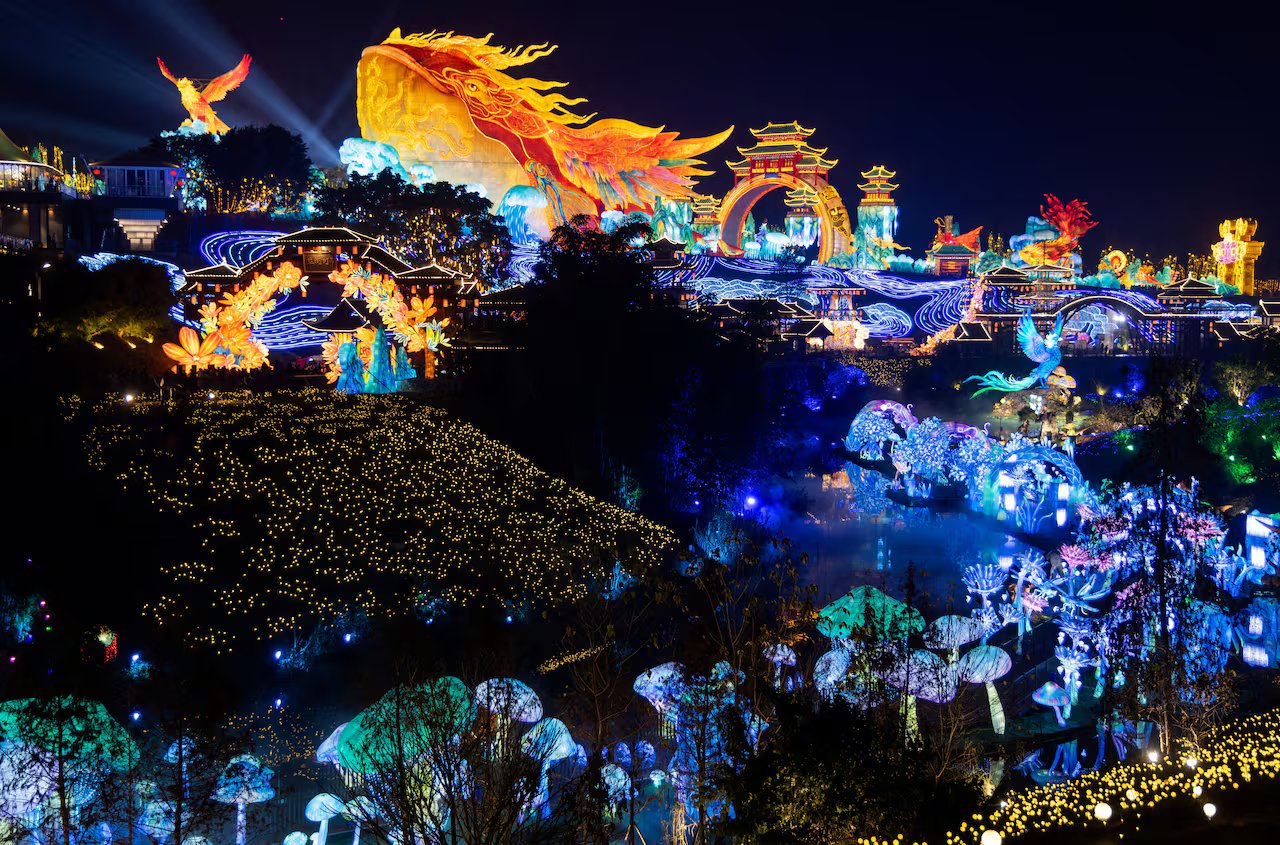 Una vista dall'alto di un parco con installazioni di lanterne giganti di diverse forme e dimensioni di animali ed edifici illuminati di notte.