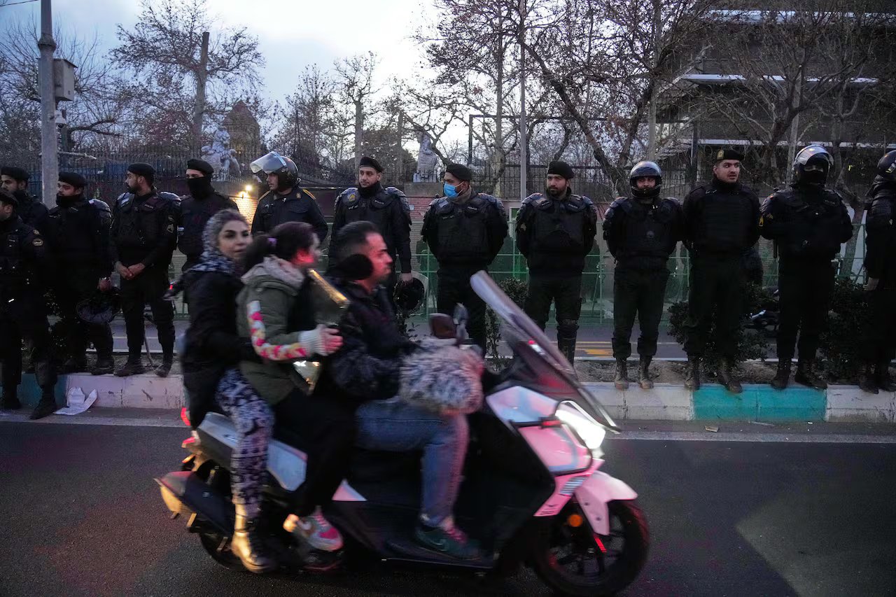 Tre persone alla guida di una motocicletta superano una fila di poliziotti.