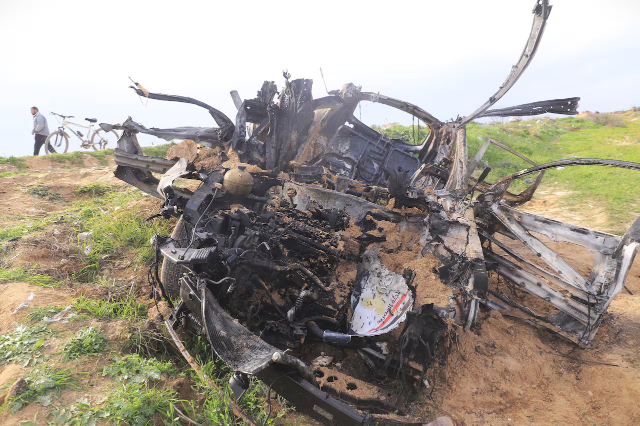 Il corpo dell'auto bombardata giace nella terra. Sullo sfondo si vedono un uomo e una bicicletta.