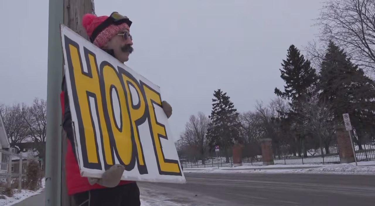 Un uomo che indossa un cappotto rosso e un cappello rosa tiene in mano un cartello con la scritta: