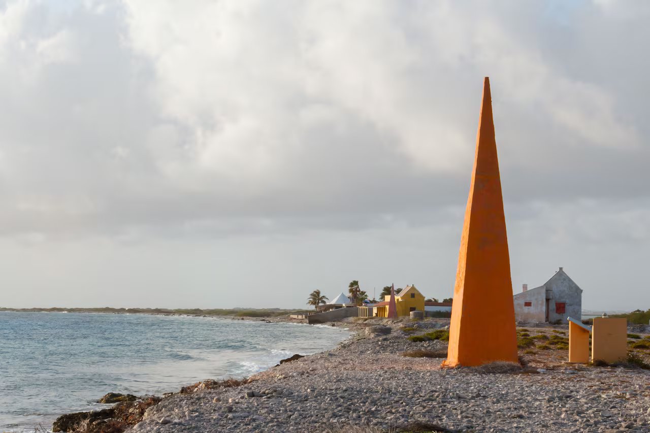 Un obelisco alto 10 metri si trova sulla spiaggia con altri edifici bassi colorati e palme in lontananza, mentre il tramonto si avvicina.