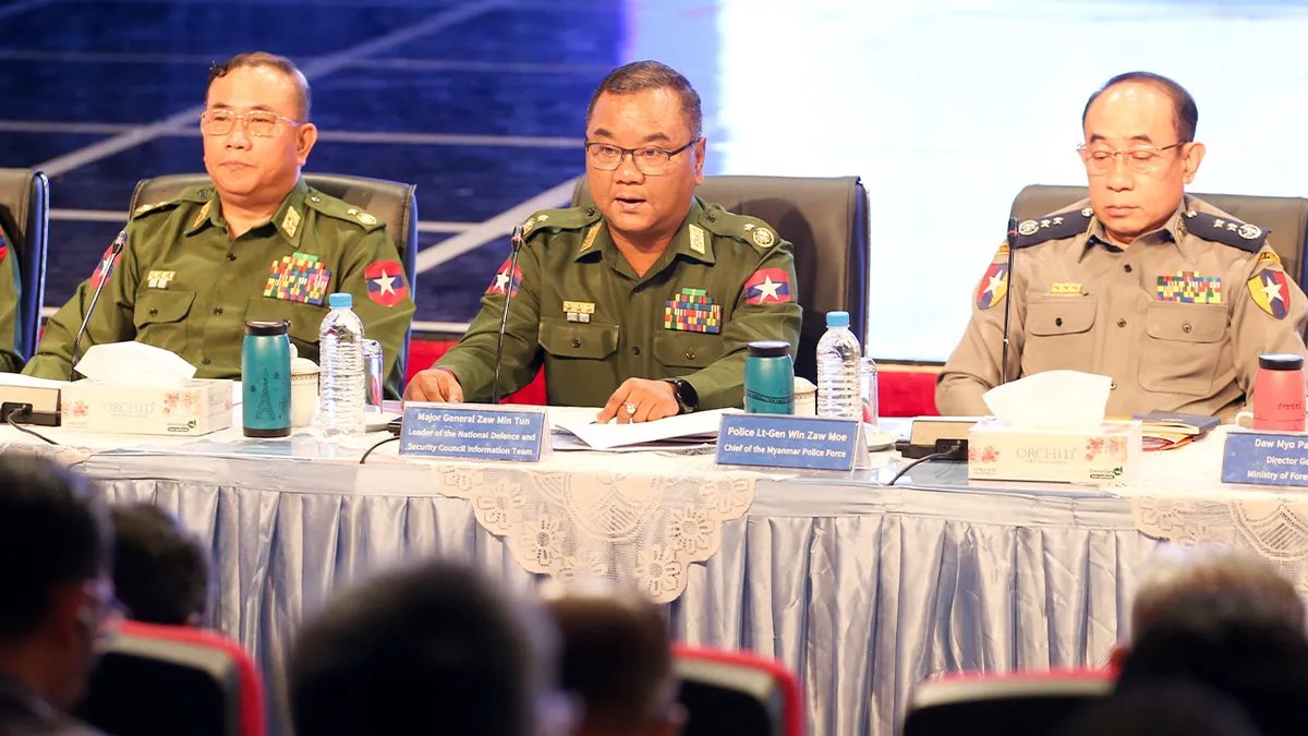 Un funzionario governativo parla sul palco durante un briefing, con i microfoni e uno sfondo alle sue spalle.