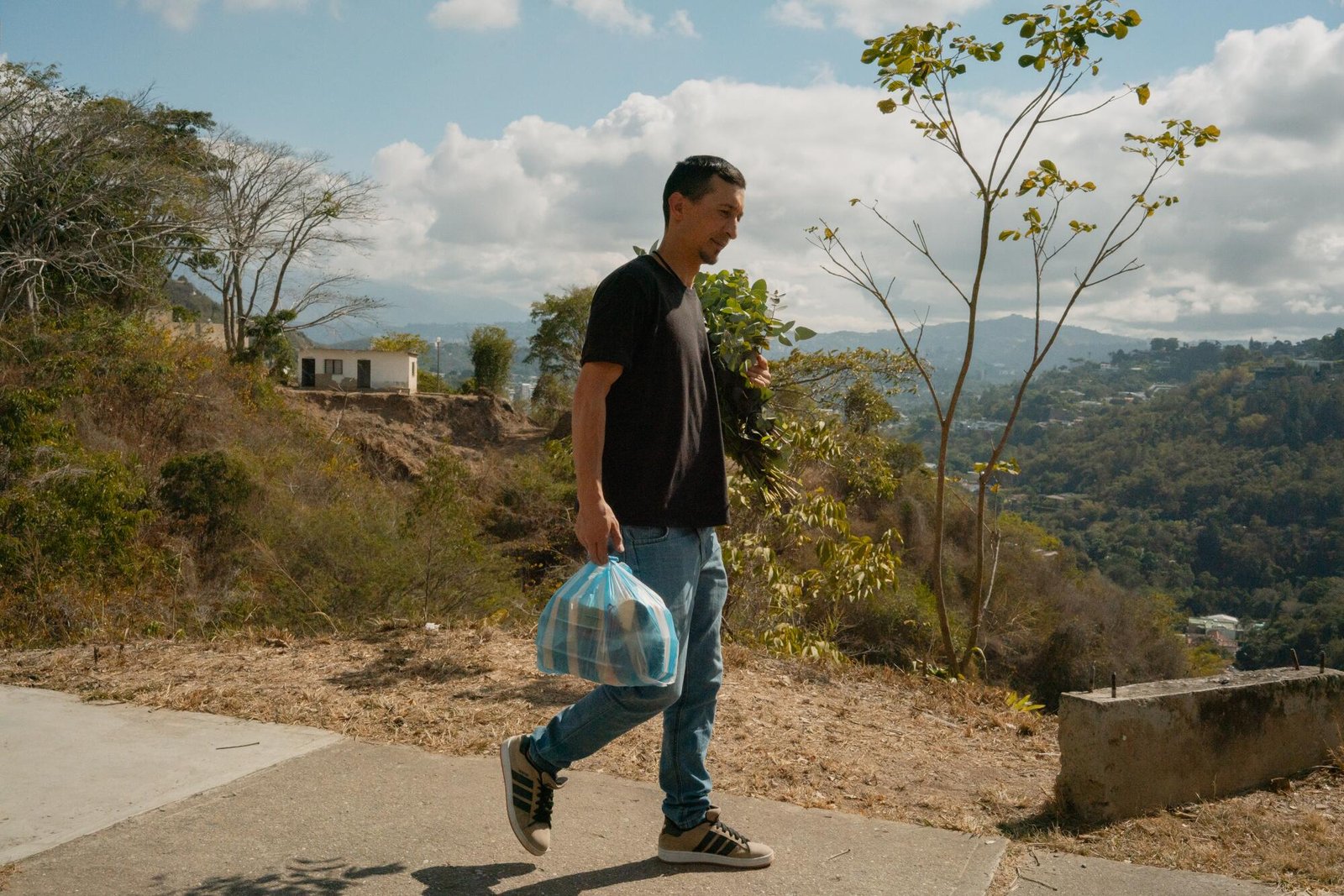 Uomo dai capelli scuri, che indossa una camicia scura e pantaloni blu, porta con sé un sacchetto di plastica a strisce blu mentre cammina in una zona montuosa