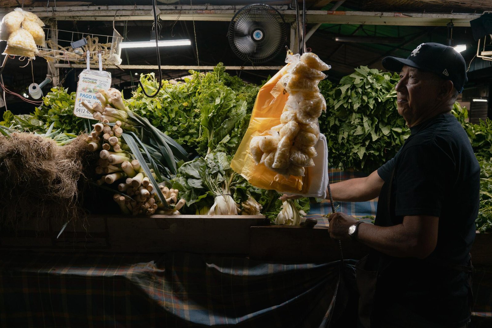 Un uomo che indossa un cappello e una camicia scuri alla sua bancarella del mercato che espone verdure, cipolle, pomodori e altri prodotti