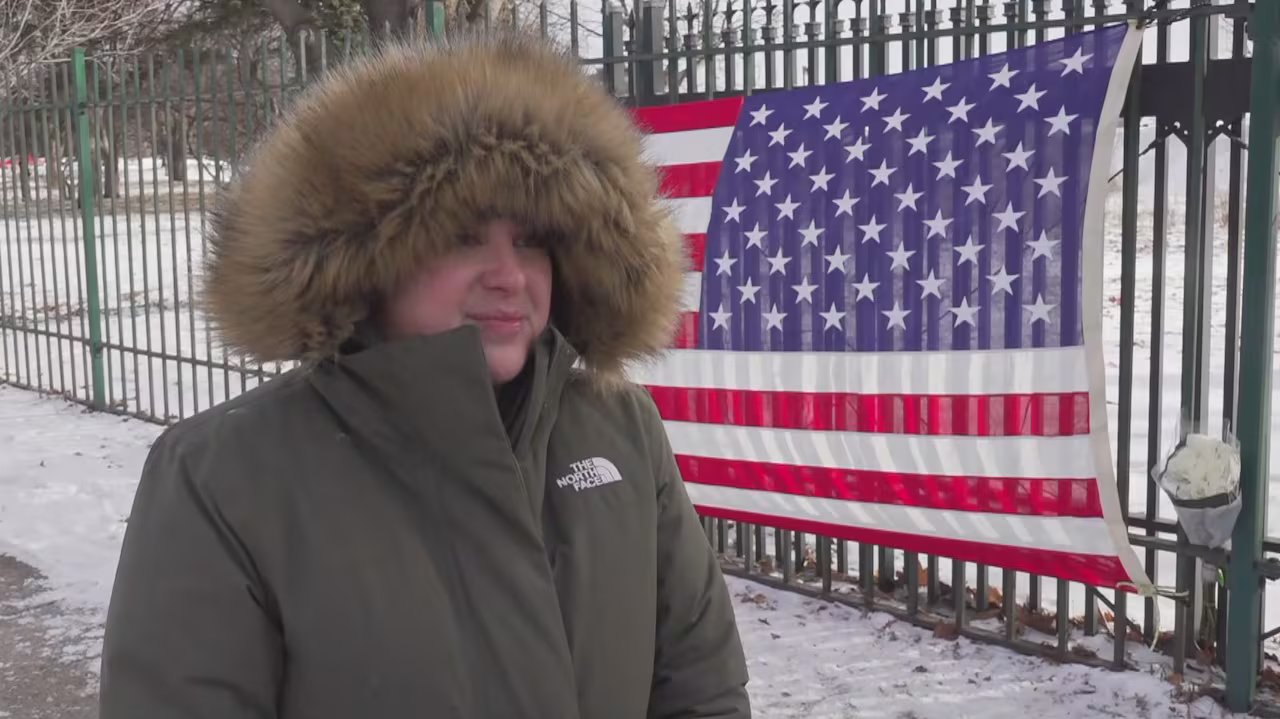 Ashley Watson indossa una giacca e si trova di fronte a una bandiera americana drappeggiata su una recinzione.