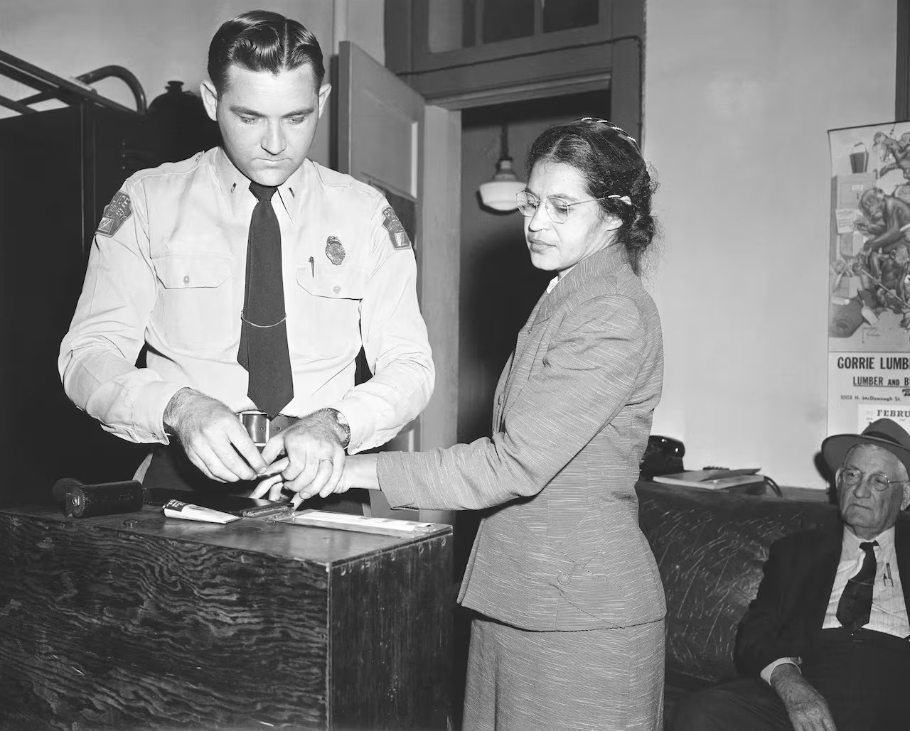 Foto in bianco e nero di una donna di colore a cui vengono prese le impronte digitali da un agente di polizia bianco