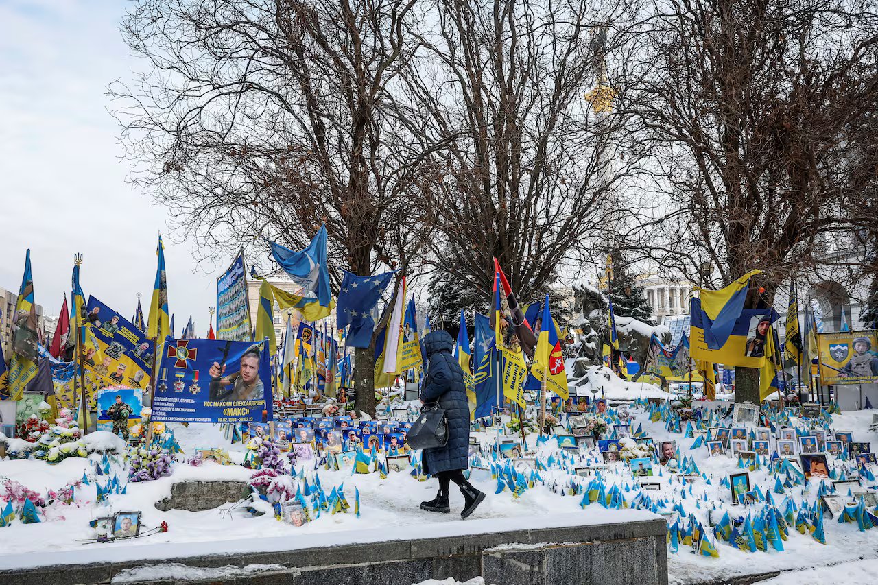 Una donna che dà le spalle alla telecamera e indossa un cappotto invernale con cappuccio visita un campo simile a un cimitero pieno di bandiere e striscioni blu e gialli.