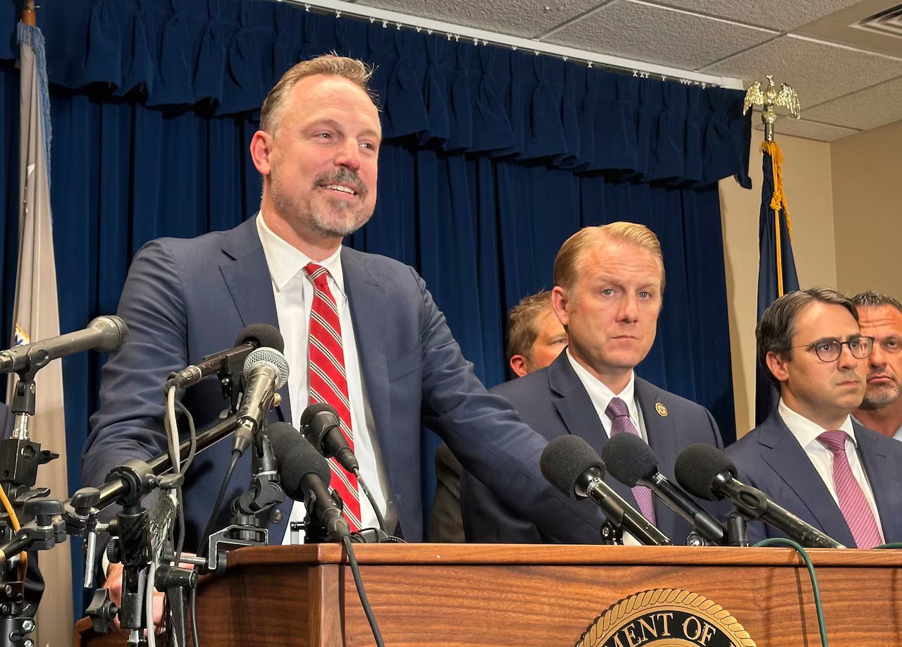 Un uomo in giacca e cravatta parla sul palco davanti a diversi microfoni. Molte persone stanno dietro di lui. 