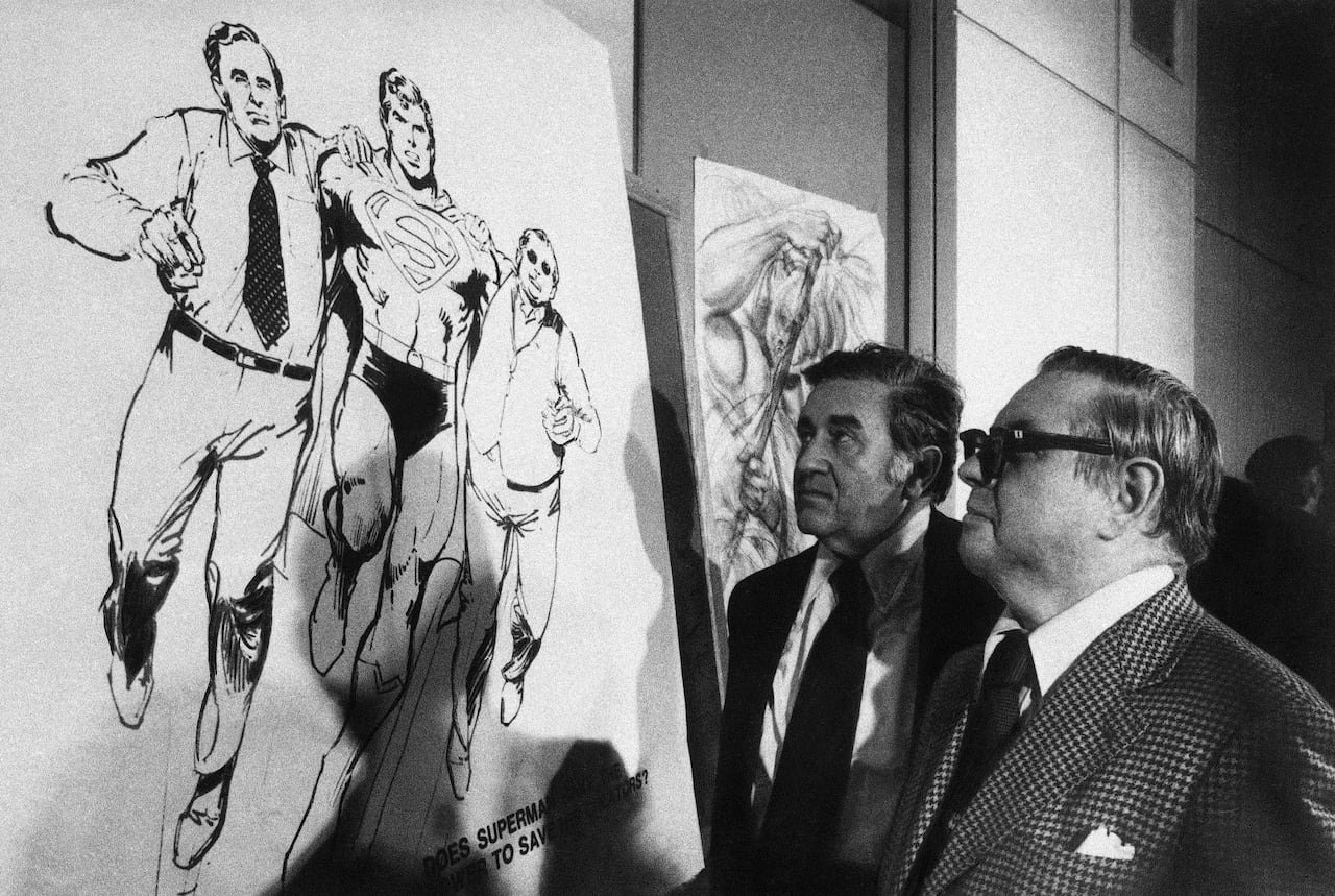 Foto in bianco e nero di due uomini in giacca e cravatta, sul lato destro dell'inquadratura, che guardano una lavagna bianca con un'illustrazione dell'eroe dei fumetti Superman che cammina con altri due uomini appoggiati sulle sue spalle.