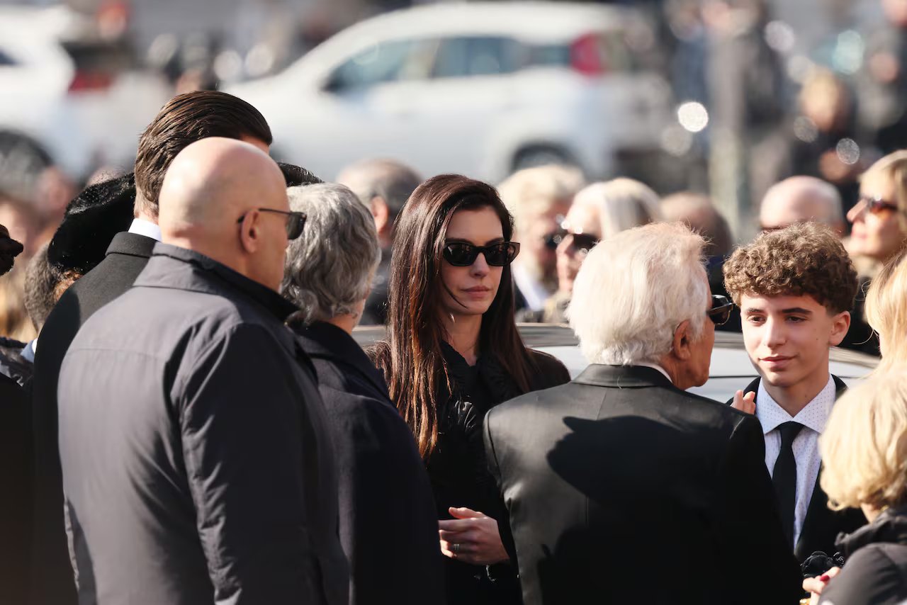Una donna che indossa occhiali da sole e abiti neri si trova all'aperto in mezzo a una folla di persone. Lei è accanto a un giovane in giacca e cravatta, e di fronte a loro ci sono uomini più anziani, anche loro in giacca e cravatta. 